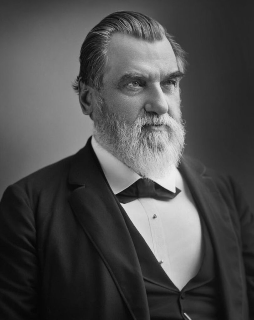 Black and white portrait of Leland Stanford. He is wearing a suit. His hair is combed back, his white beard almost falling onto his chest. He looks towards the right edge of the image.